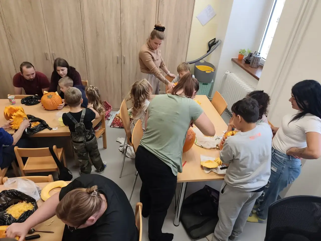 Děti i dospělí společně dlabou dýně u stolů ve třídě